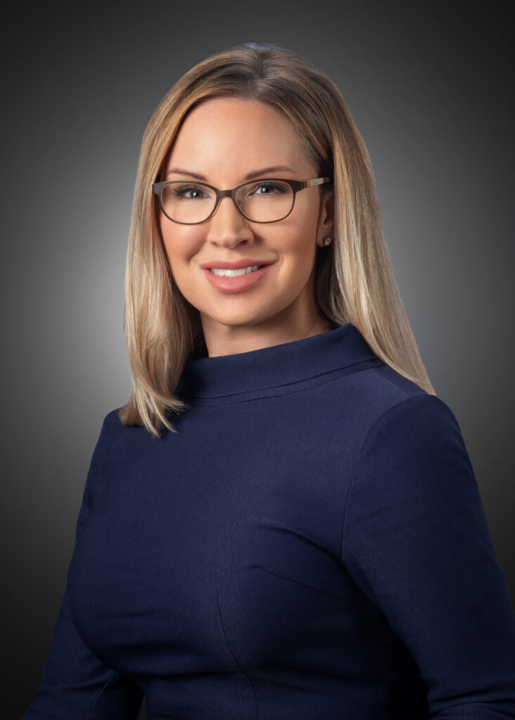 Studio headshot of a woman wearing glasses and a navy dress, smiling slightly against a dark gray background.
