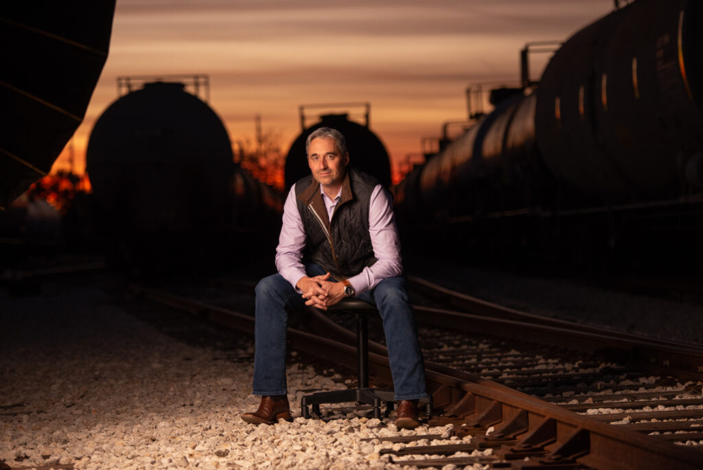 Environmental company executive seated on a stool in a rail yard at sunset, with blurred rail cars in the background.