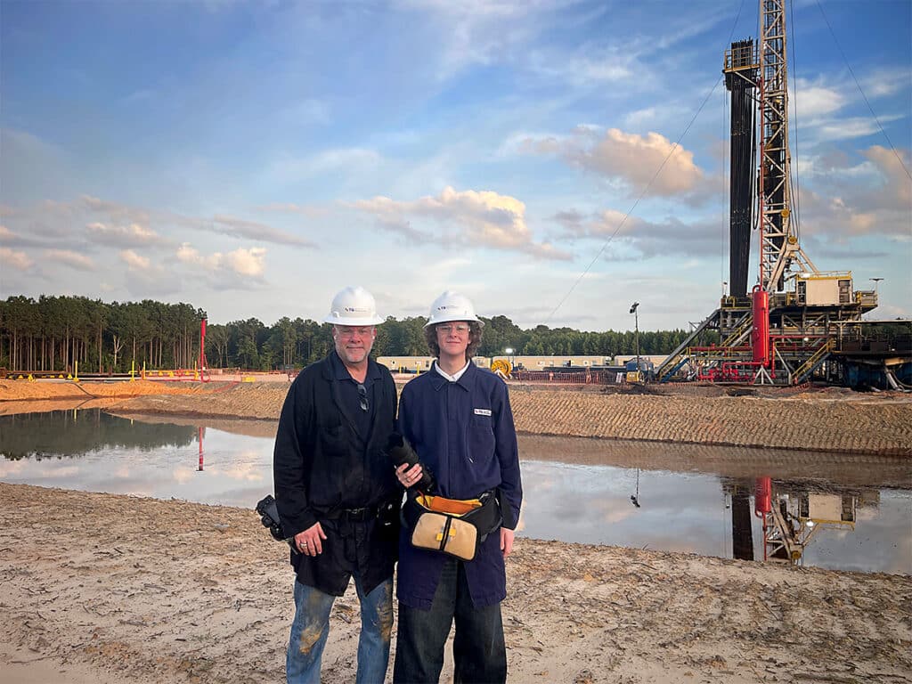 Photographer Aric with his son Gavin on a Texas oil and gas drilling site, wearing hard hats at sunset. Photographed on an iPhone by a security escort during a three-day industrial photography assignment.