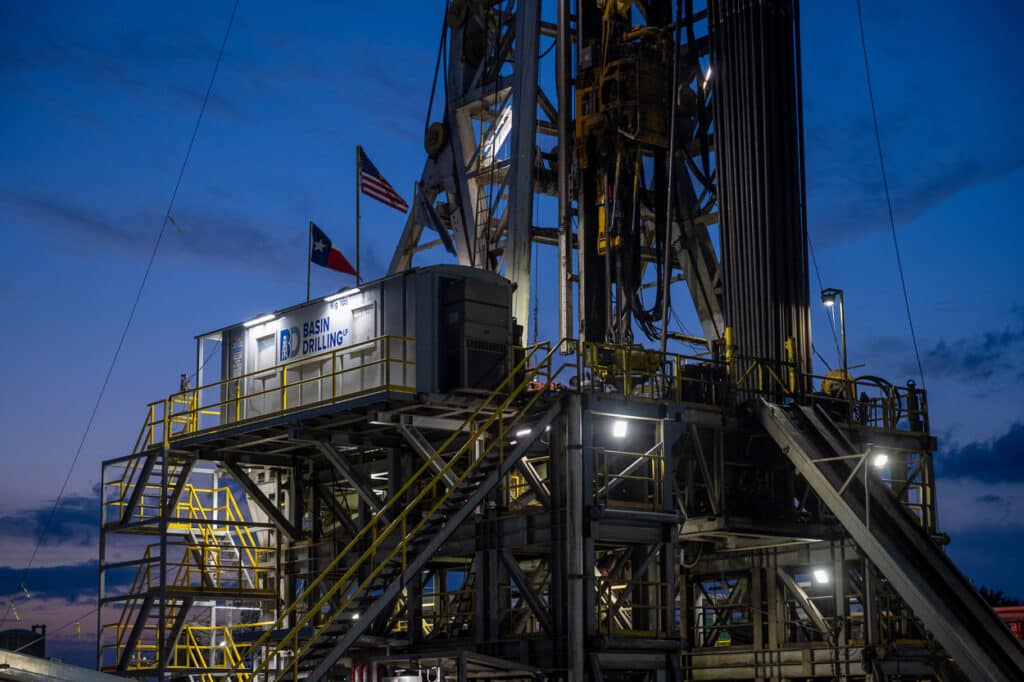 Twilight view of Basin Drilling rig in East Texas, fully lit with safety lighting and American and Texas flags visible, oil and gas safety report photography.