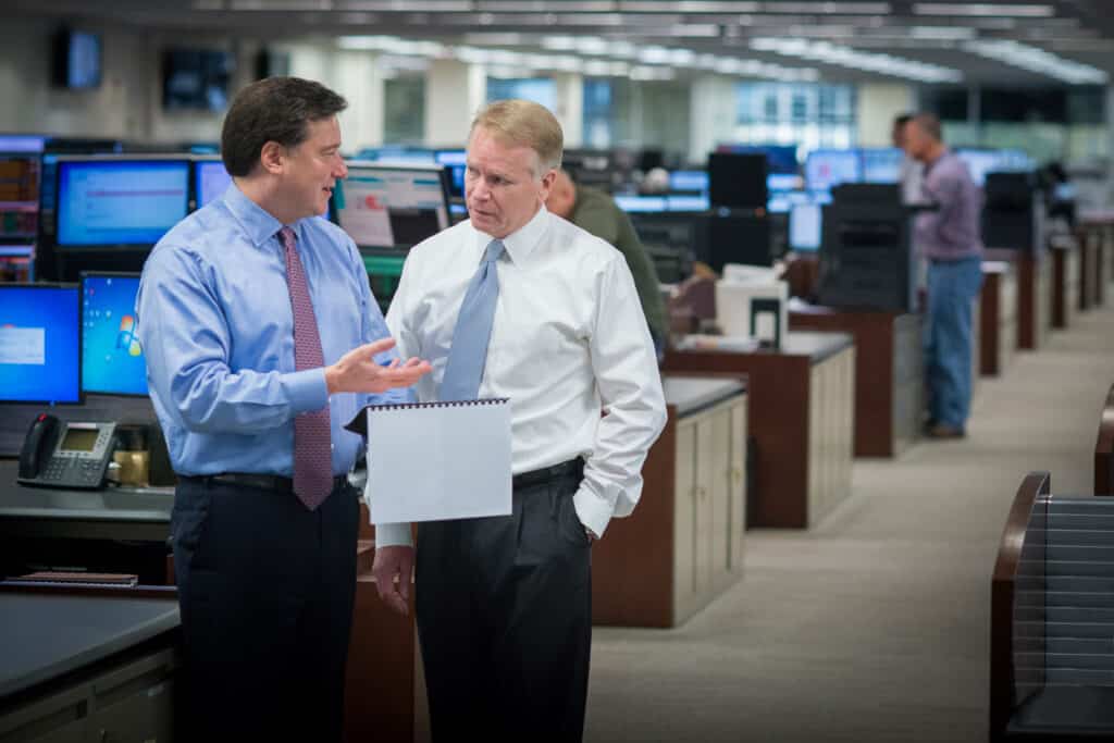 Two business professionals in conversation on a trading floor, surrounded by computer monitors and workstations, captured for a corporate annual report.