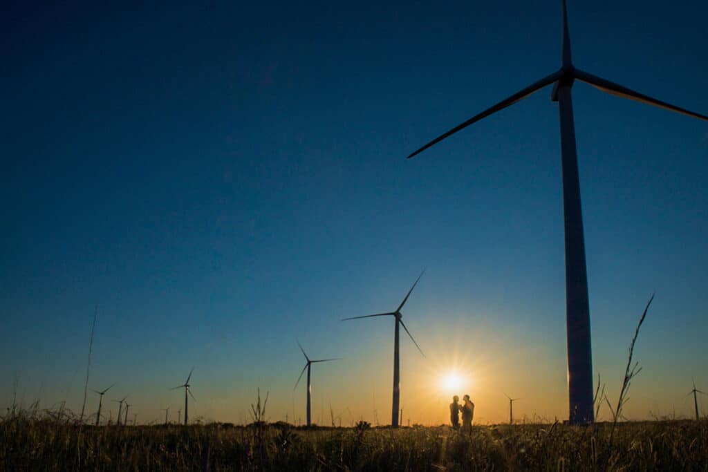 Two people silhouetted at sunset beneath large wind turbines in a grassy field, captured for an annual report with natural lighting and wide-angle composition.