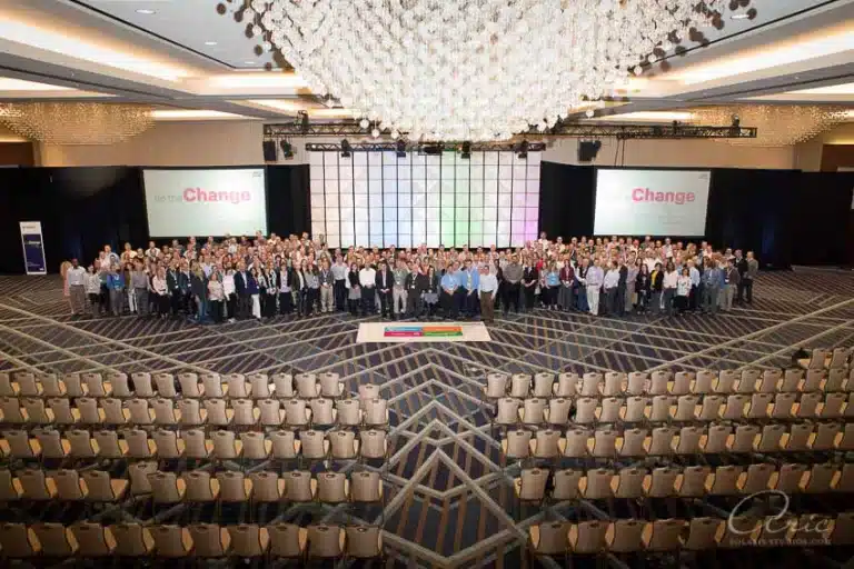 Large group photo of conference attendees on a stage in a ballroom, with rows of empty chairs in the foreground and presentation screens in the background.