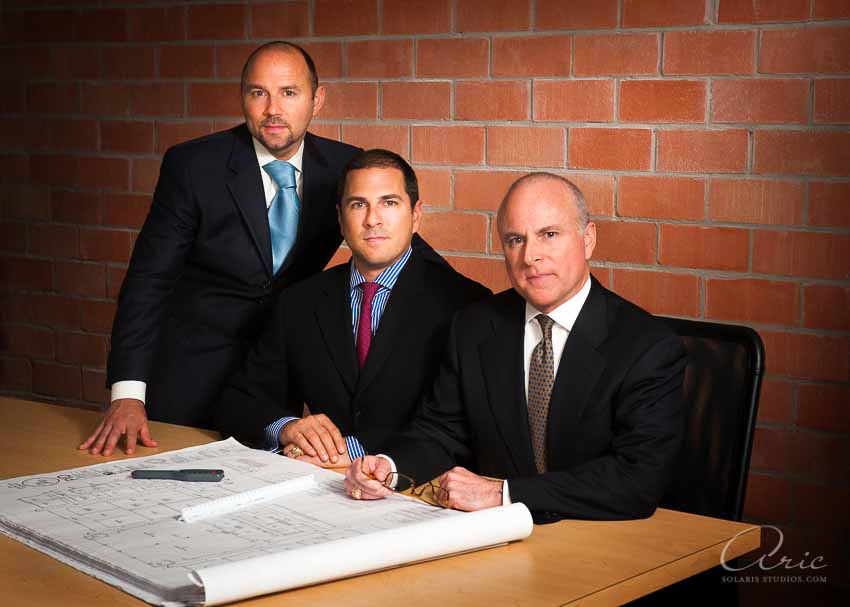 Group portrait of three executives in suits gathered around a conference table with architectural blueprints, against a brick wall.