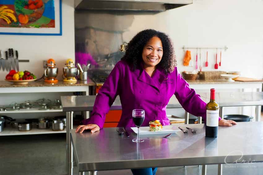 Smiling chef in a purple jacket leaning on a stainless-steel counter in a commercial kitchen, with plated food and a glass of wine.