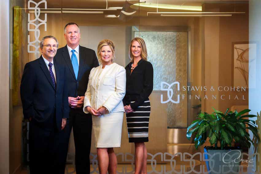 Group portrait of four professionals standing behind branded glass in an office lobby for Stavis & Cohen Financial.