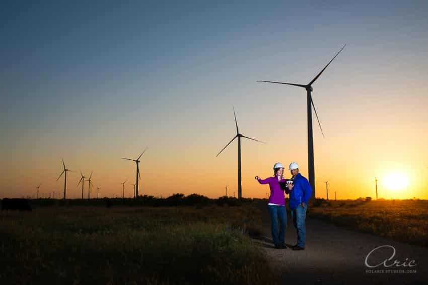 Two workers wearing hard hats standing on a road at a wind farm at sunset, with multiple wind turbines in the background.