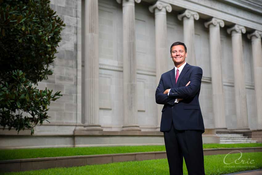Executive in a suit standing outdoors with arms crossed in front of a large building with stone columns for Annual Report Photography.