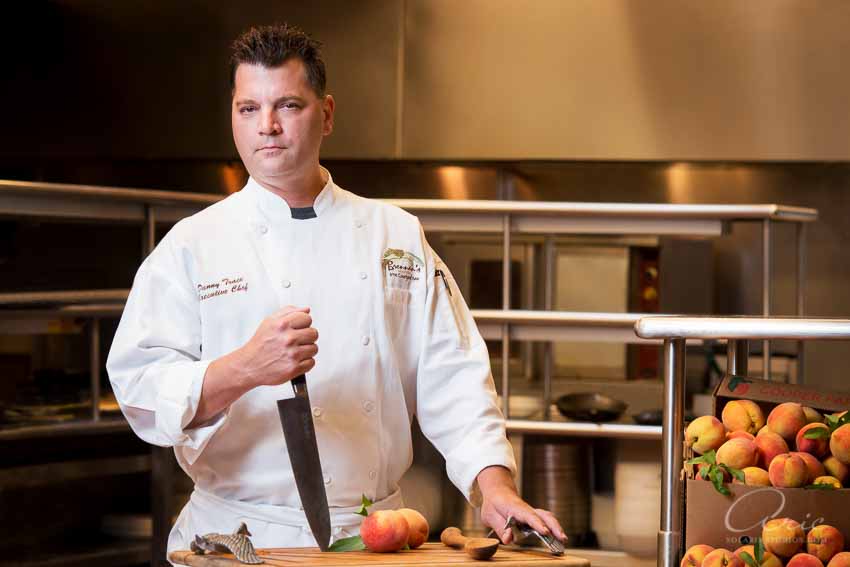 Portrait of the executive chef at Brennan’s of Houston standing in the kitchen in a white chef coat, holding a chef’s knife beside fresh peaches on a prep table