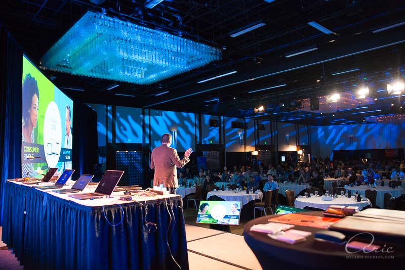 Business conference keynote speaker presenting on stage with presentation screens and audience seated at round tables during a corporate event