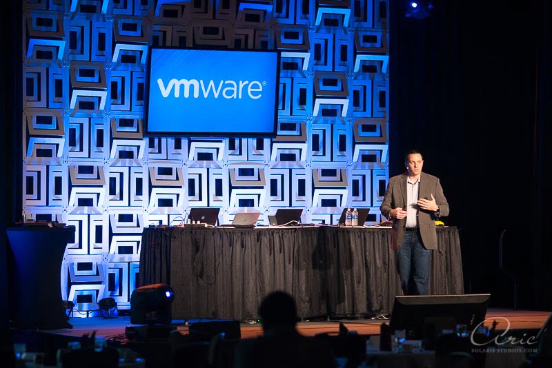 VMware technology conference keynote speaker presenting on stage with branded backdrop and presentation equipment during a corporate event