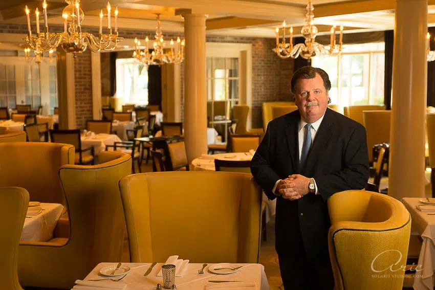 Executive in a dark suit standing in an upscale restaurant dining room with chandeliers and yellow chairs.