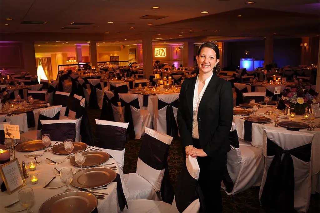 Environmental professional headshot of event coordinator in elegant banquet hall photographed with balanced off-camera lighting and wide-angle composition for corporate branding portrait