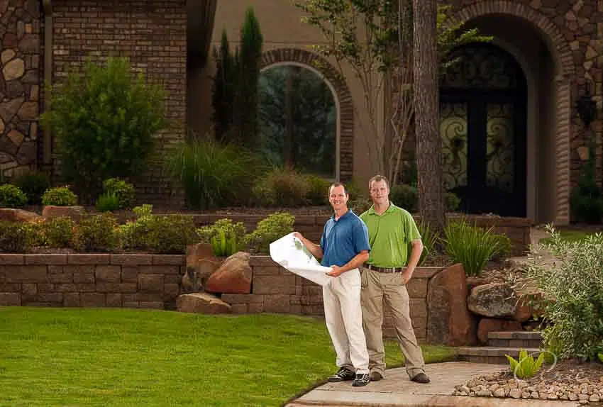 Environmental corporate team portrait of landscape design professionals reviewing site plans outside luxury residential property photographed with natural daylight and wide environmental composition