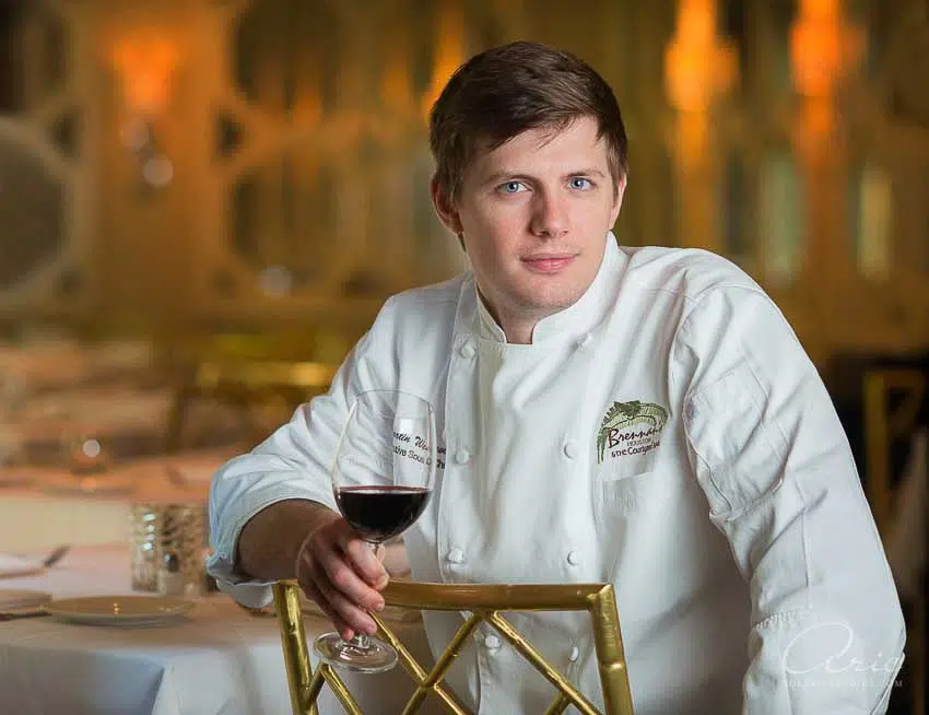 Environmental executive portrait of professional chef holding wine glass in upscale restaurant photographed with soft ambient lighting and shallow depth of field for hospitality branding
