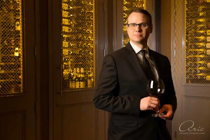 Environmental executive portrait of wine director holding glass in upscale wine cellar photographed with warm ambient lighting and shallow depth of field for hospitality leadership branding