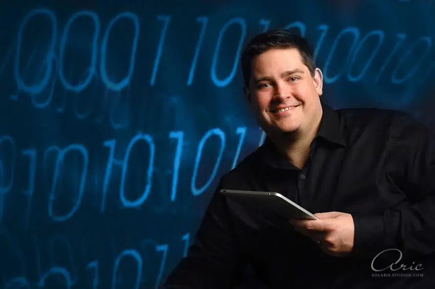 Professional business headshot of technology executive holding tablet photographed with directional studio lighting and digital binary background for corporate leadership profile