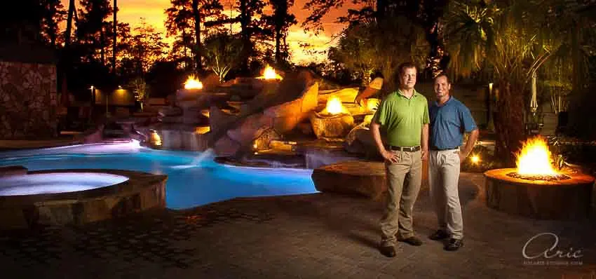 Environmental corporate team portrait of landscape design professionals standing beside luxury outdoor pool and fire features photographed with dramatic evening lighting for project branding