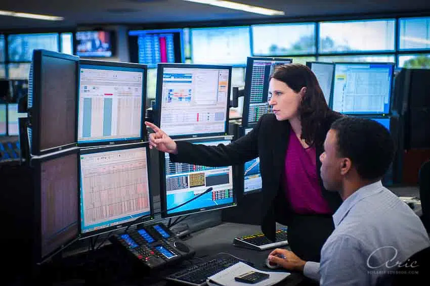 Two professionals in a control room reviewing data on multiple computer monitors, with one person pointing at a screen.