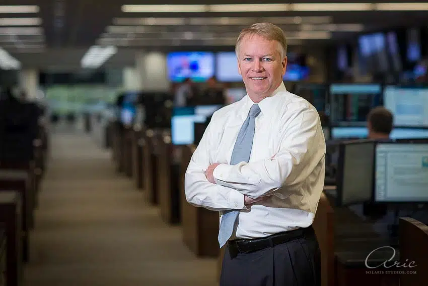 Environmental executive portrait of finance professional in trading operations center photographed with balanced office lighting and shallow depth of field for corporate leadership branding