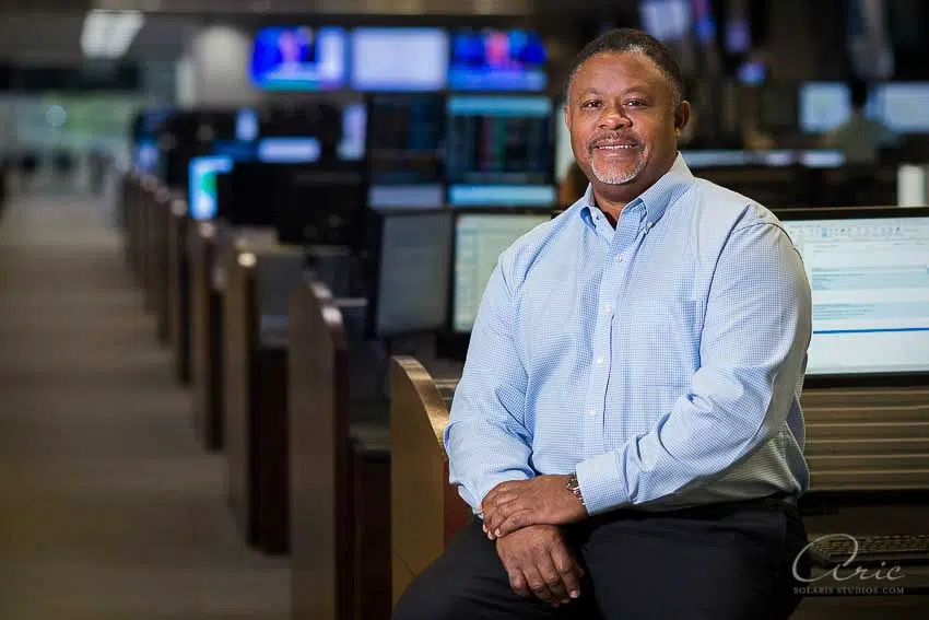 Environmental executive portrait of finance operations leader seated in trading center photographed with balanced office lighting and shallow depth of field for corporate leadership branding