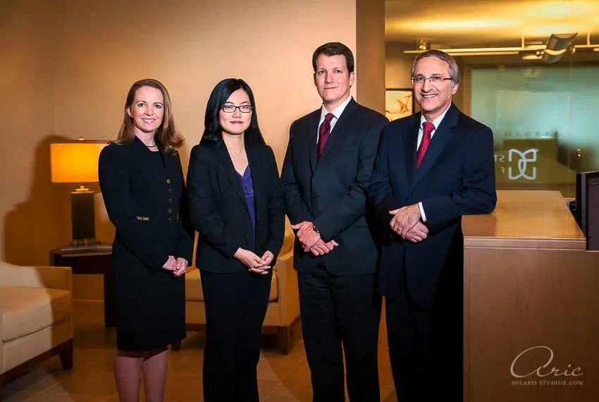 Corporate leadership team group portrait of four executives photographed in modern office reception area with balanced off-camera flash lighting and environmental branding backdrop for professional company profile