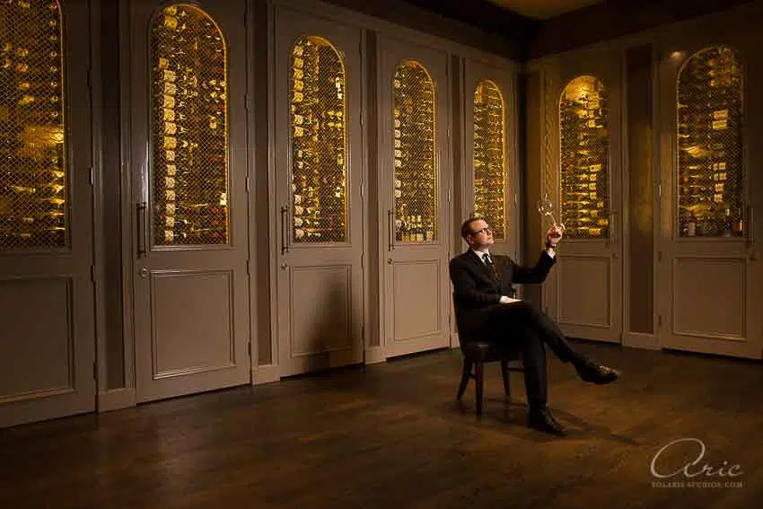 Environmental executive portrait of wine director evaluating glass in luxury wine cellar photographed with warm ambient lighting and wide architectural composition for hospitality branding