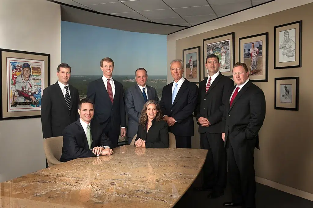 Executive leadership team portrait in a boardroom with a conference table, framed artwork on the walls, and a city view in the background.