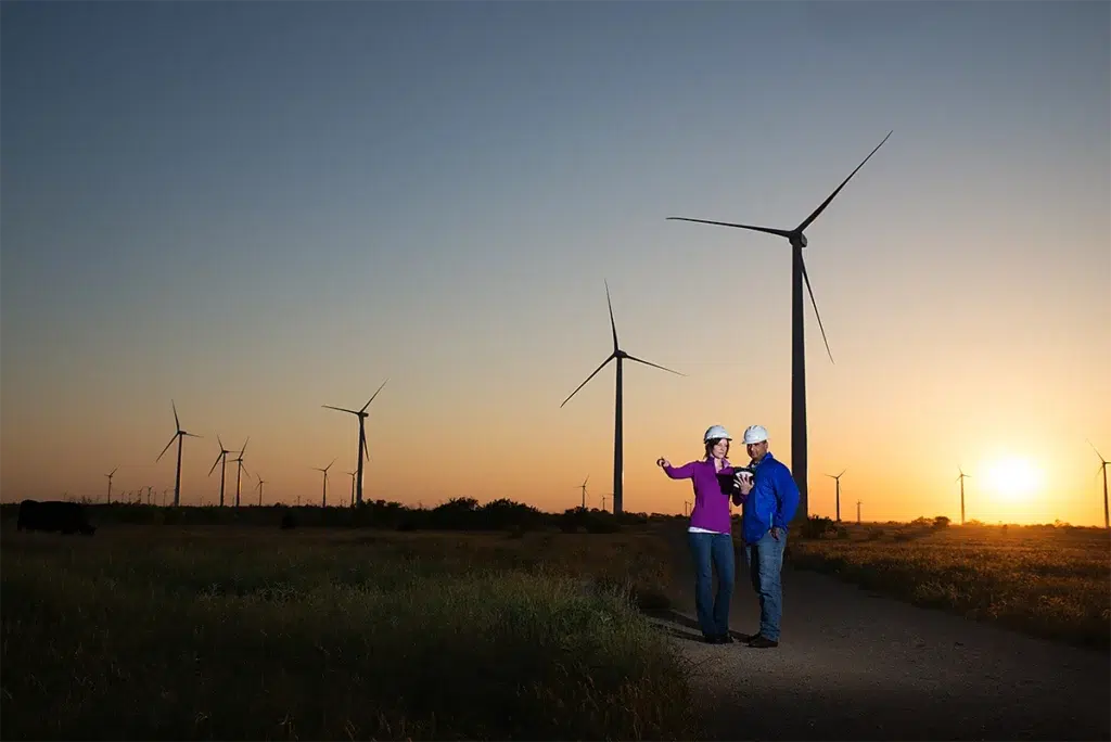 Environmental corporate portrait of renewable energy engineers reviewing project at wind farm during sunset photographed with dramatic natural backlighting and wide landscape composition