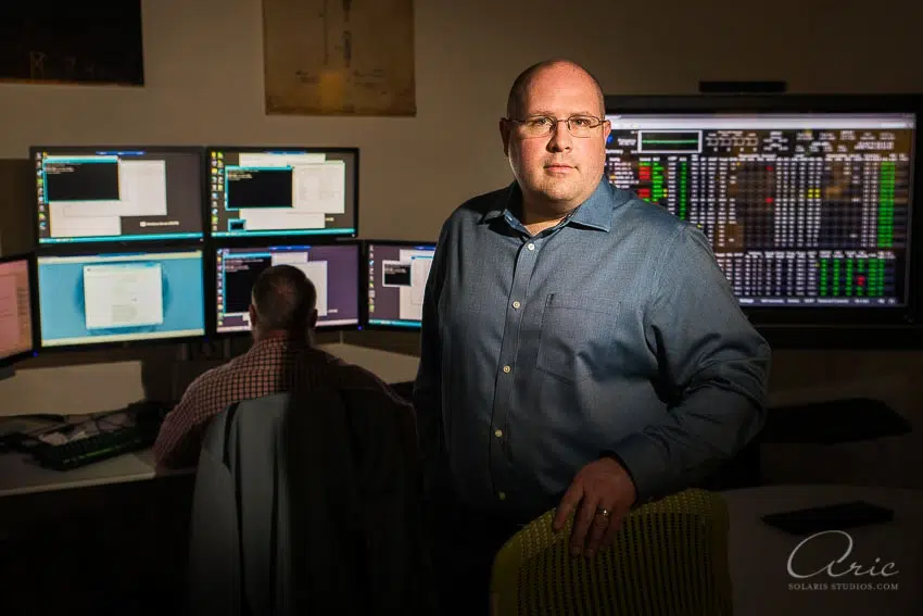 Environmental executive portrait of technology operations manager in data monitoring center photographed with dramatic directional lighting and multi-screen workstation background for leadership branding