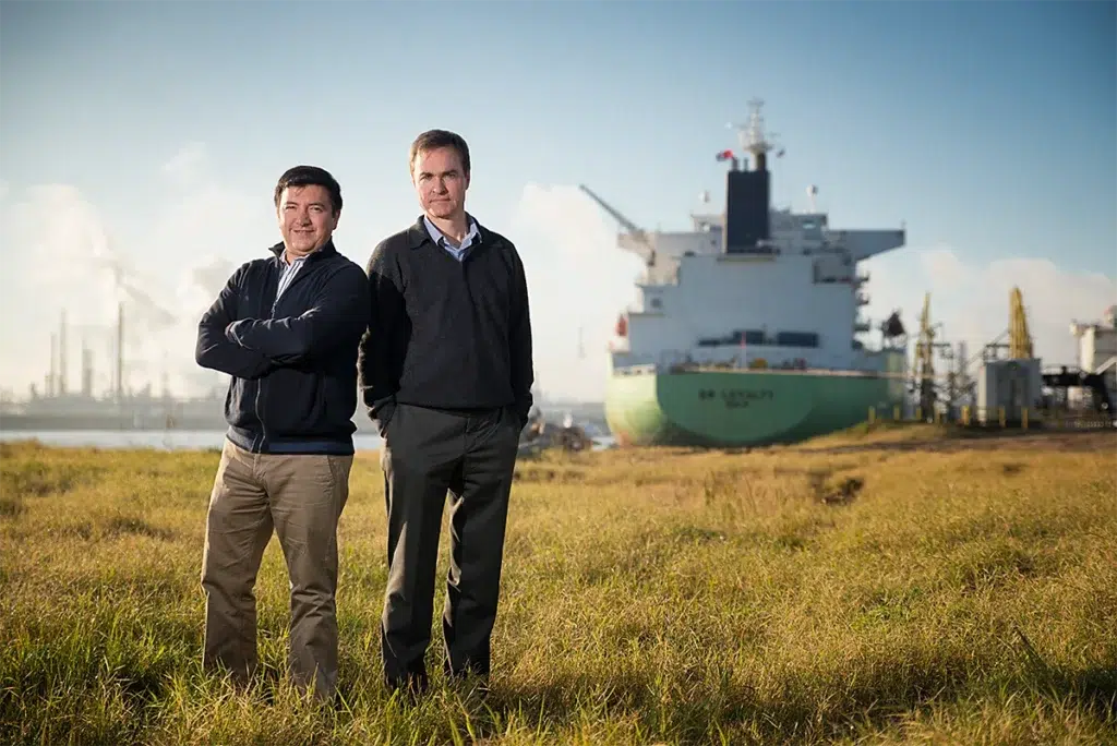 Two business leaders standing in a grassy field near an industrial port, with a cargo ship in the background.