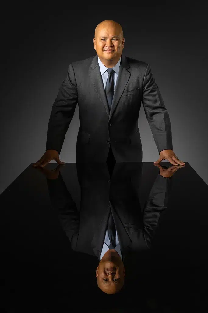 Studio executive portrait of business leader photographed with dramatic directional lighting and reflective conference table composition against dark background for corporate leadership branding