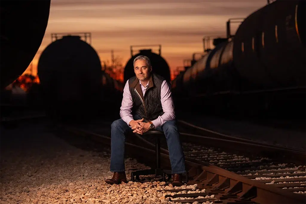 Environmental executive portrait of business leader seated on rail track between tanker cars at sunset photographed with dramatic off-camera lighting and shallow depth of field for executive branding