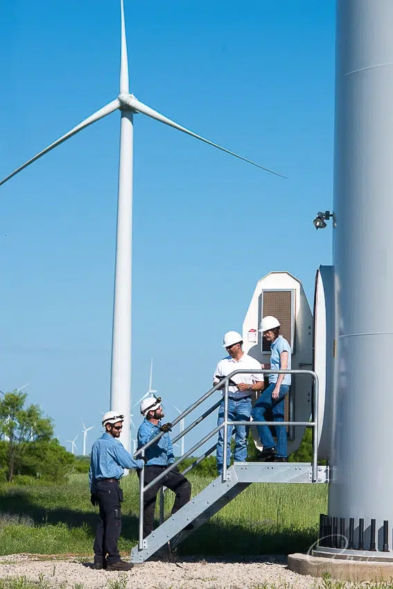 Environmental corporate team portrait of wind energy engineers at turbine site photographed with natural daylight and wide environmental composition for corporate leadership branding