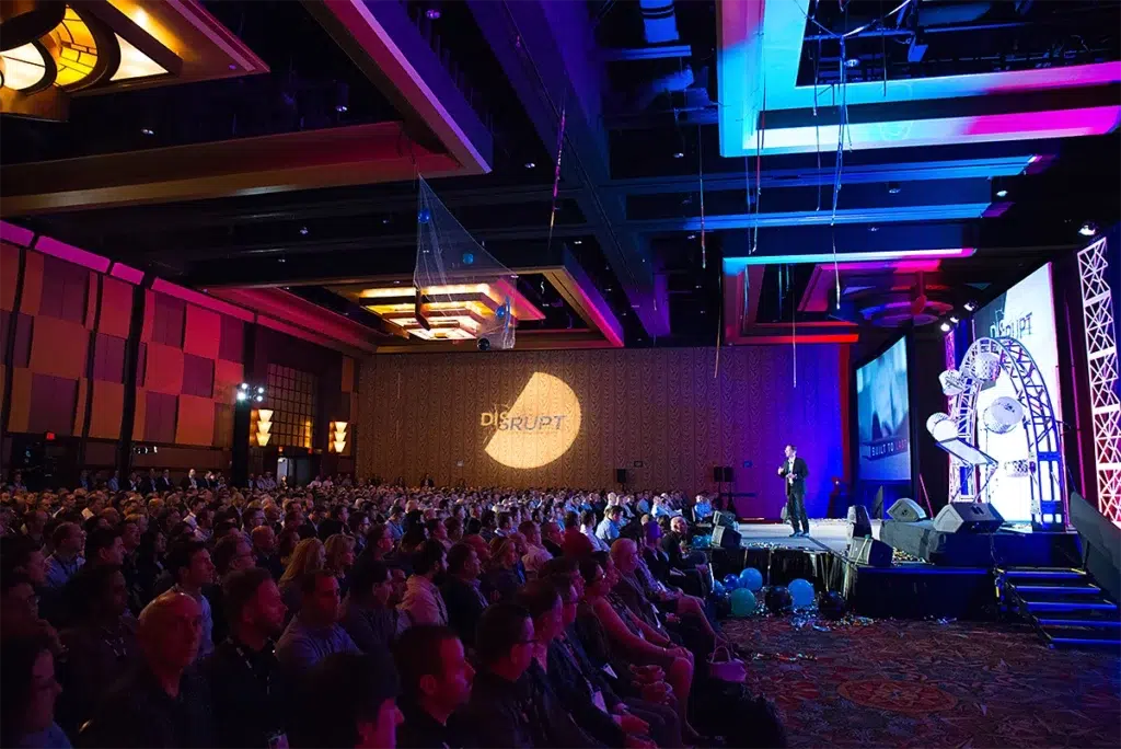 Large corporate conference audience watching a keynote presentation on stage during a business event with colorful stage lighting and branded visuals