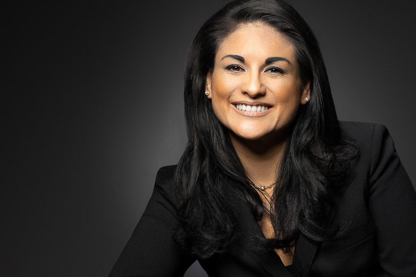 Studio headshot of a smiling woman in a black blazer against a dark background.