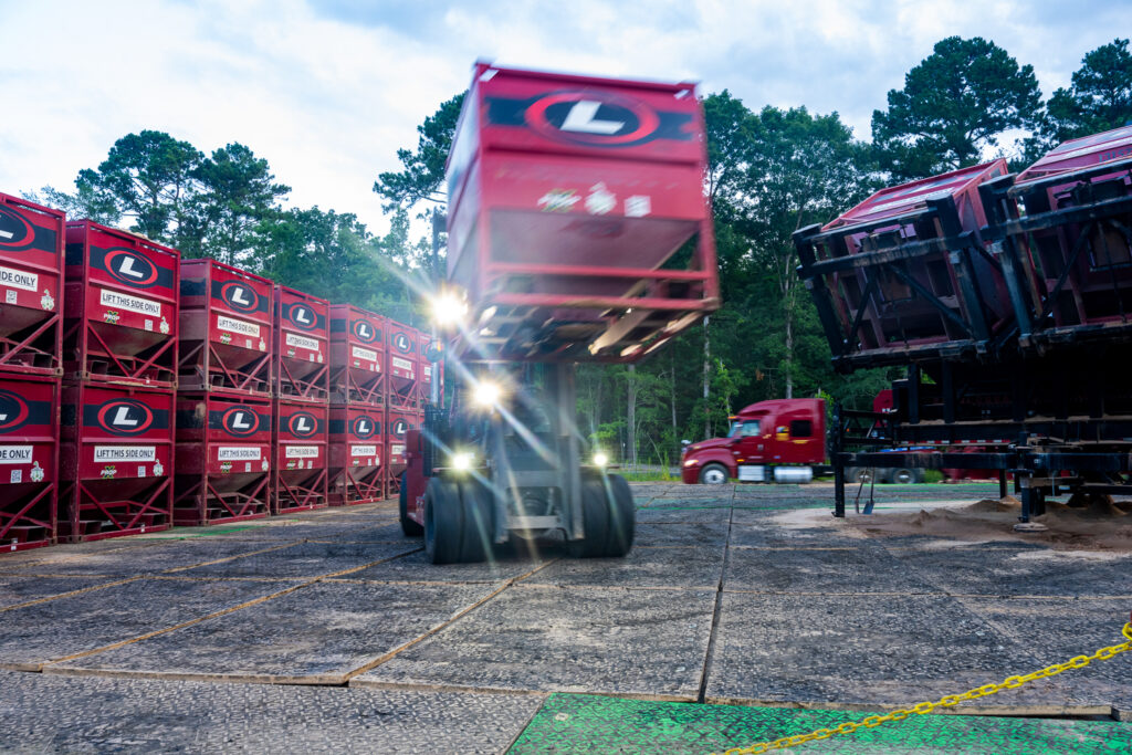 Forklift moving a large red sand container at a fracking site, with stacked containers and an 18-wheeler in the background.