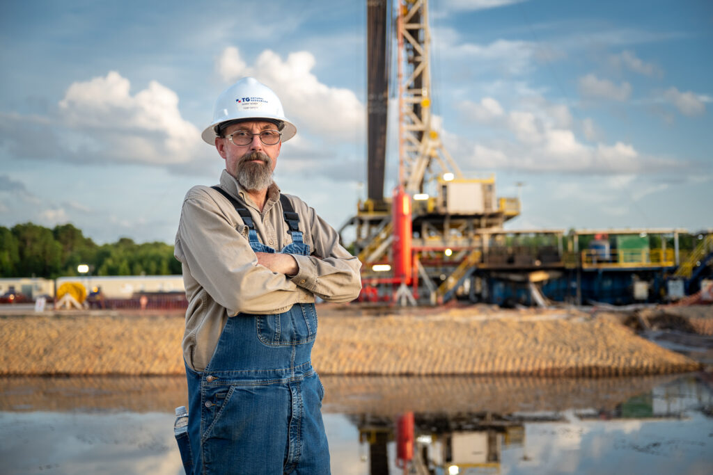 Worker wearing a hard hat and safety glasses standing with arms crossed at an active drilling site, with a rig in the background.