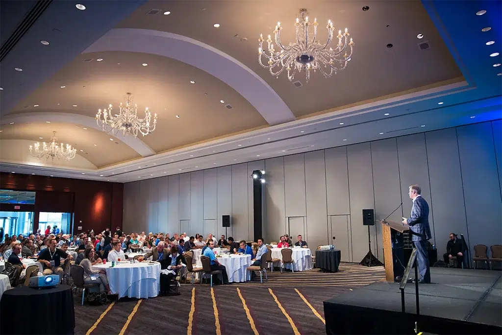 Wide corporate event photograph of a speaker at a podium addressing a seated audience in a ballroom with chandeliers and high vaulted ceilings in Houston.