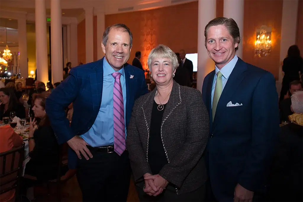 Three business leaders posing together during a formal corporate gala event inside an elegant ballroom with warm ambient lighting