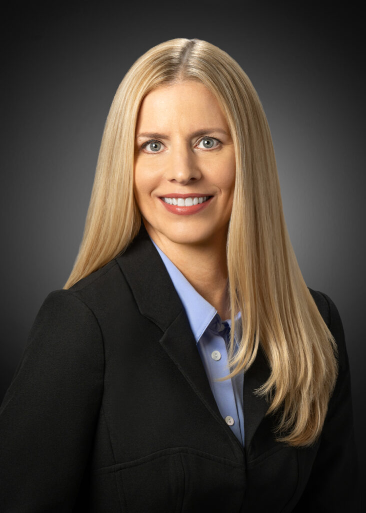 Professional studio headshot of female executive with long blonde hair wearing black blazer and blue blouse photographed with soft studio lighting and dark gray gradient background for corporate branding and LinkedIn profile