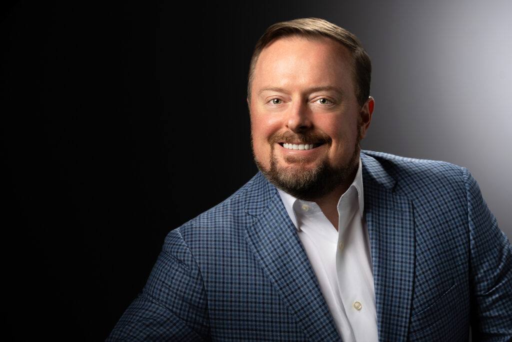 Professional studio headshot of male executive in blue patterned jacket photographed with dramatic directional studio lighting and split gradient background for corporate branding and LinkedIn profile