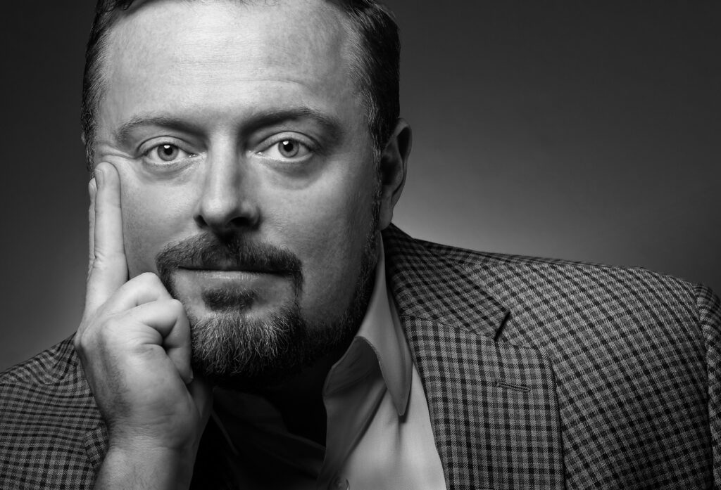 Black and white studio headshot of male executive resting his hand on his face photographed with dramatic studio lighting and neutral gradient background for professional corporate branding. For Headshot Photography Portfolio
