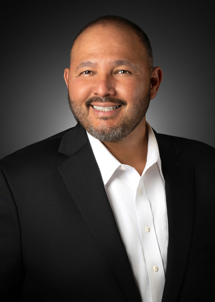 Professional studio headshot of smiling male executive in black suit photographed with soft directional studio lighting and dark gradient background for corporate branding and LinkedIn profile