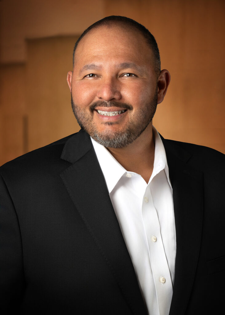Professional studio headshot of smiling male executive in black suit photographed with soft studio lighting and warm gradient background for corporate profile and LinkedIn branding