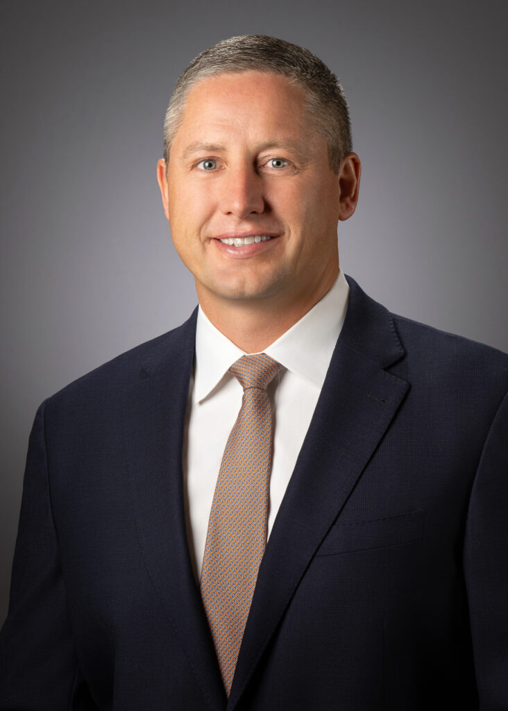 Professional studio headshot of male executive wearing navy suit and orange tie photographed with soft studio lighting and neutral gray background for corporate branding and LinkedIn profile. For Headshot Photography Portfolio