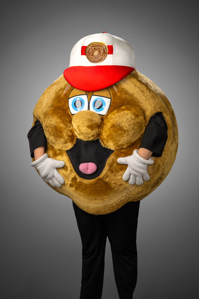 Studio portrait of donut mascot character wearing branded red and white Shipley Do-Nuts hat photographed against gray studio background for commercial branding and promotional marketing