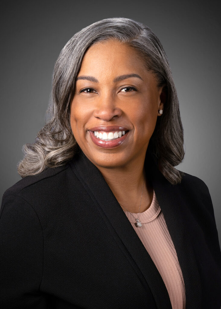 Professional studio headshot of female executive with gray hair wearing black blazer photographed with soft studio lighting and dark gray gradient background for corporate branding and LinkedIn profile