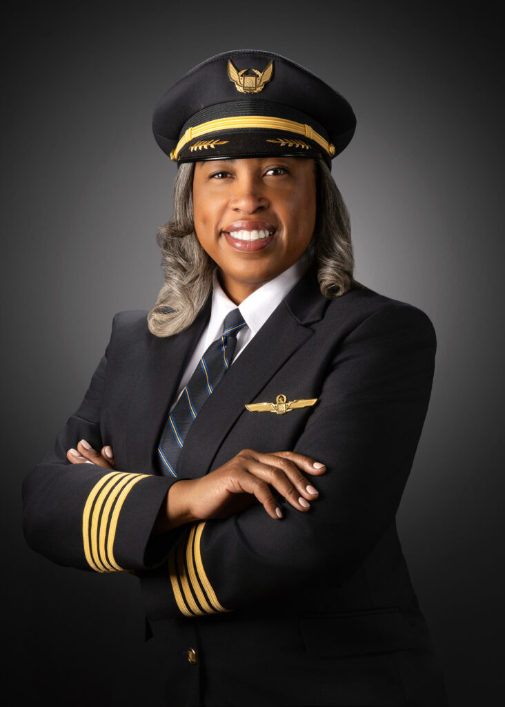 Professional studio portrait of female airline pilot in uniform with arms crossed photographed with soft studio lighting and dark gray gradient background for executive branding and leadership profile
