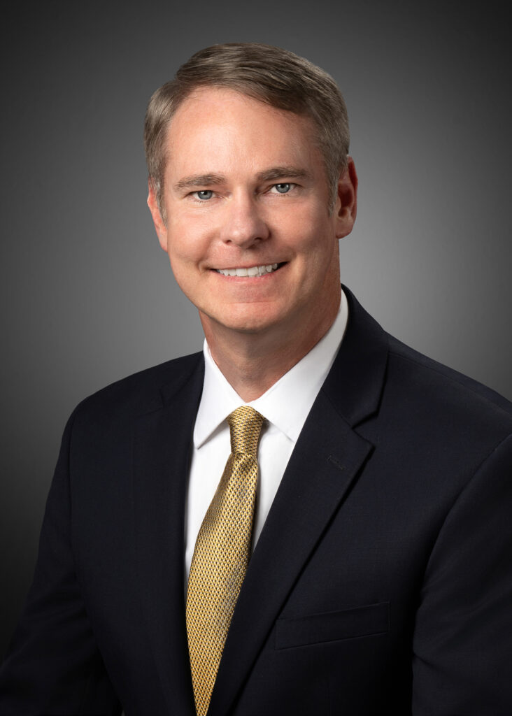 Professional studio headshot of male executive wearing navy suit and gold tie photographed with soft studio lighting and neutral gray background for corporate branding and LinkedIn profile. For Headshot Photography Portfolio